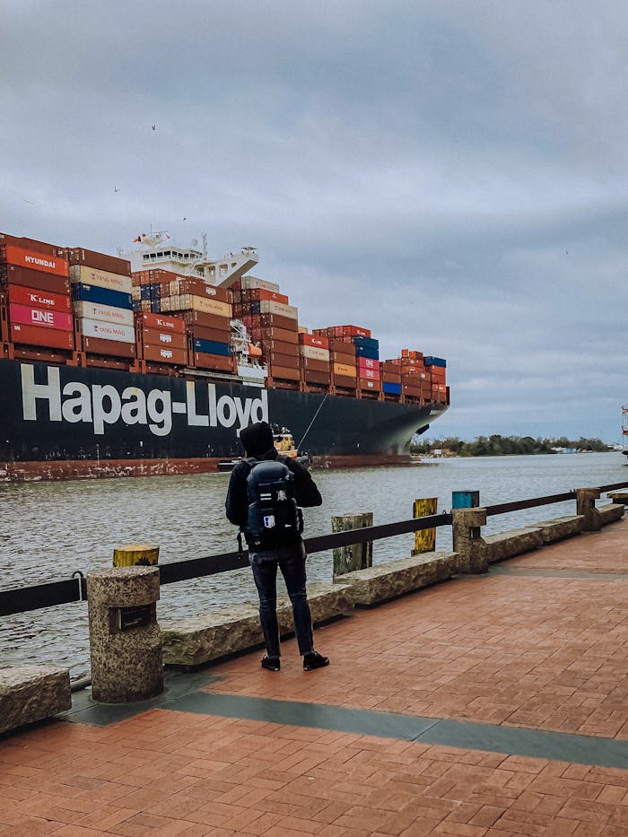 about-us A backpacker stands by a harbor, observing a large cargo ship loaded with containers.